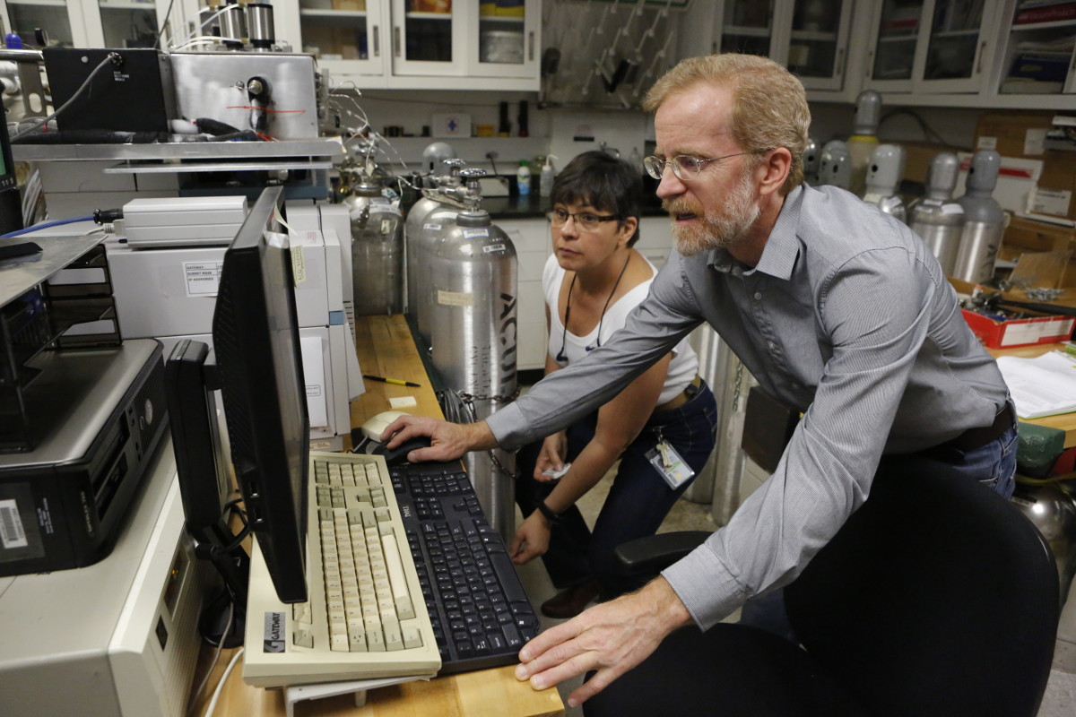 Photo of a man and woman in a scientific lab in front of a computer screen and keyboard. Both are looking at the computer screen, and additional equipment including large cylindrical flasks are in the background.